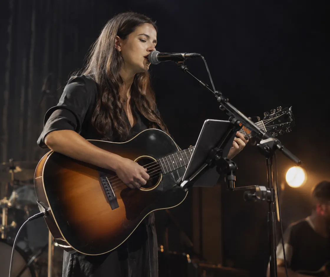 A singer-songwriter on stage, playing acoustic guitar and reading her set from a tablet mounted on the mic stand